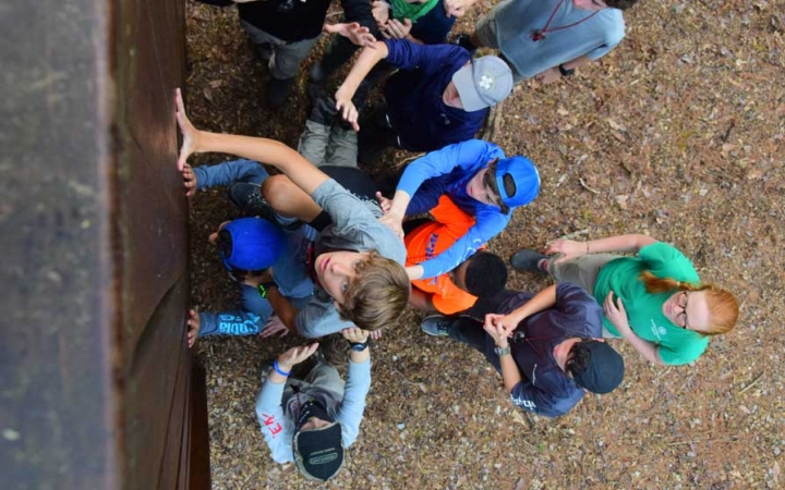 From the top of an obstacle, the camera looks down at a group of students helping another up.